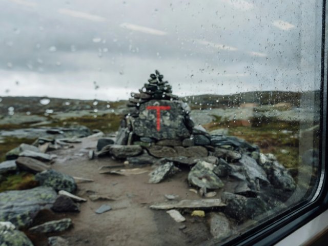 Shallow-focus view through a rain-speckled railway carriage window toward a red-T painted stone trail cairn at Finse