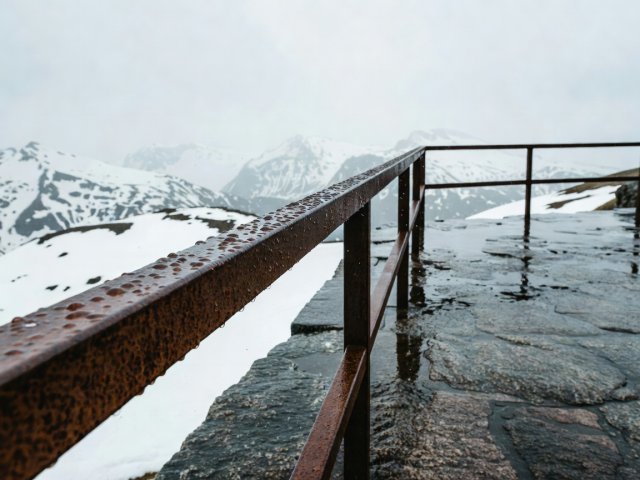 Close-up of rain beads on a Corten steel viewpoint railing with blurred mountain snowfields in the background